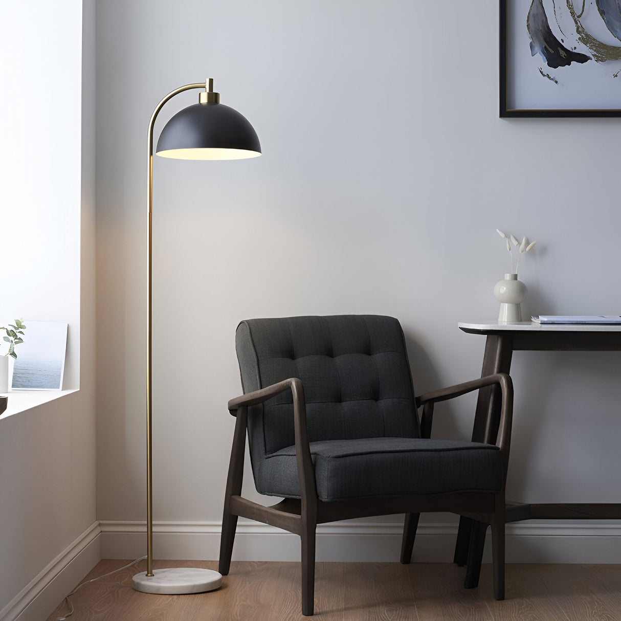 Endon Ashmont floor lamp with arched antique brass stem, matt black dome shade and marble base, beside a dark tufted armchair in a living room corner.