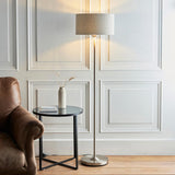 Endon Brenley floor lamp in chrome with a natural linen drum shade, lit beside a leather armchair and black side table in a panelled living room.