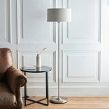 Endon Brenley floor lamp in chrome with a natural linen drum shade, styled beside a leather armchair and black round side table in a living room.