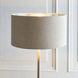 Endon Brenley floor lamp shown lit with a natural linen drum shade, metallic inner and slim chrome stem against a panelled off-white wall.