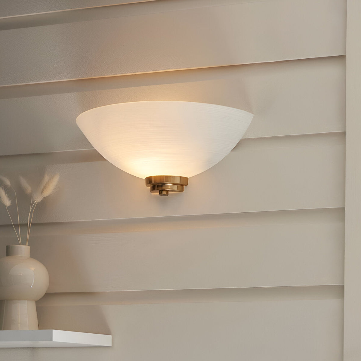 Endon Carlton wall light with matt white half-bowl glass shade and brushed aged brass fitting, illuminated on a pale panelled wall above a shelf.