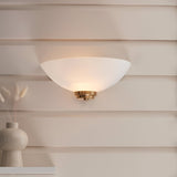 Endon Carlton wall light with a matt white glass half-bowl shade and brushed aged brass fitting, illuminated on a cream panelled wall above a vase.