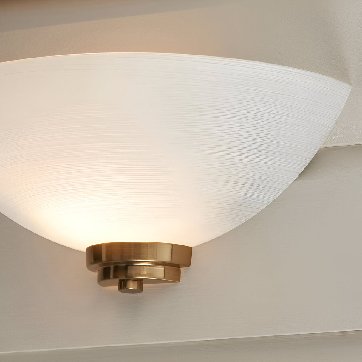 Endon Carlton wall light illuminated, with a half-bowl matt white ribbed glass shade and brushed aged brass backplate mounted on a pale wall.