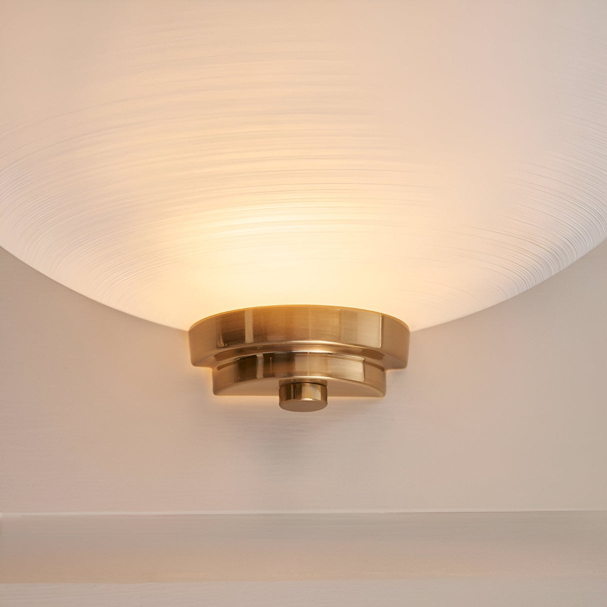 Endon Carlton wall light with a matt white ribbed glass half-shade on a brushed aged brass backplate, shown lit and mounted on a pale wall.