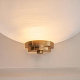 Endon Carlton wall light with a matt white ribbed glass half-shade on a brushed aged brass backplate, shown lit and mounted on a pale wall.