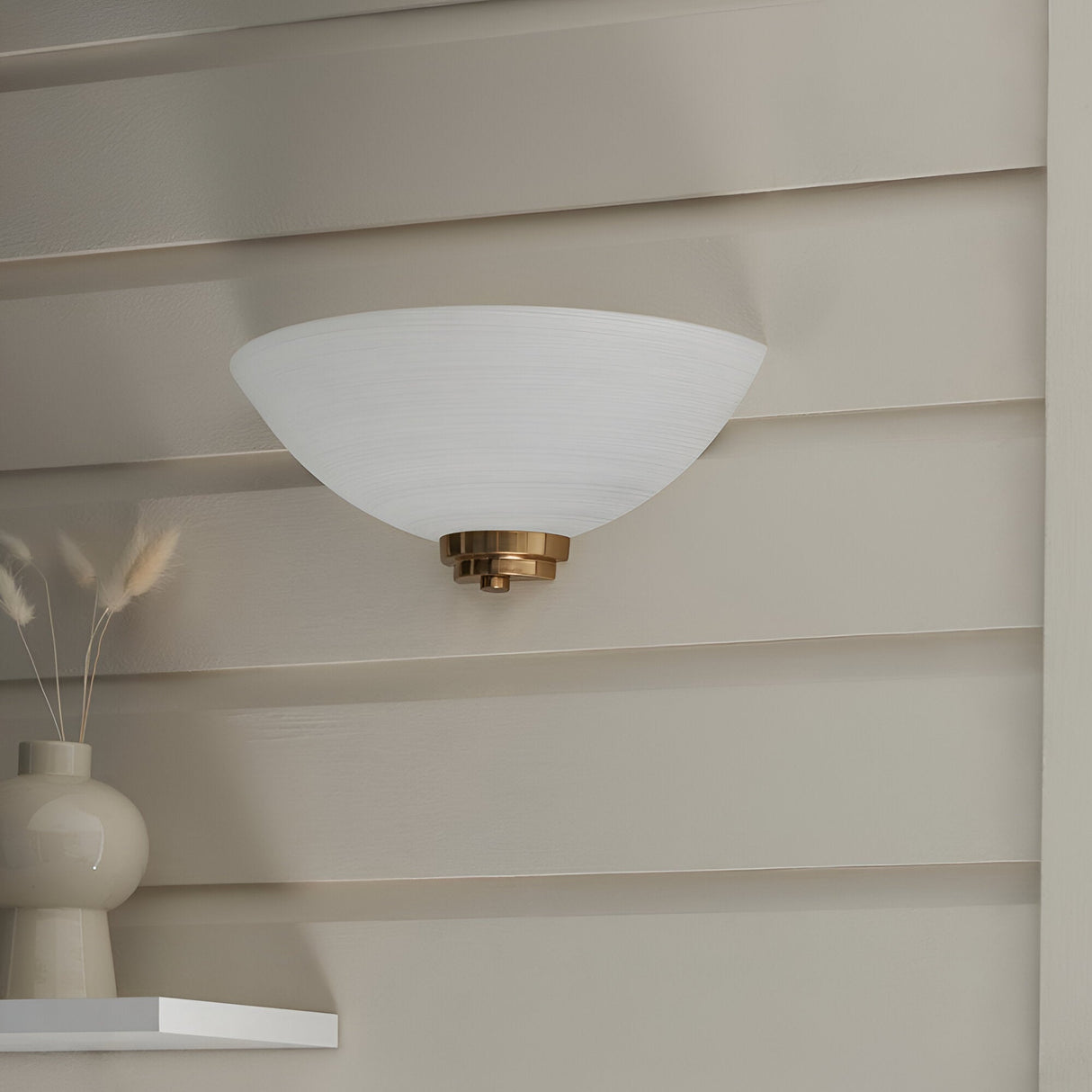 Endon Carlton wall light with matt white ribbed glass dome shade and brushed aged brass fitting, mounted on a panelled wall above dried grasses.
