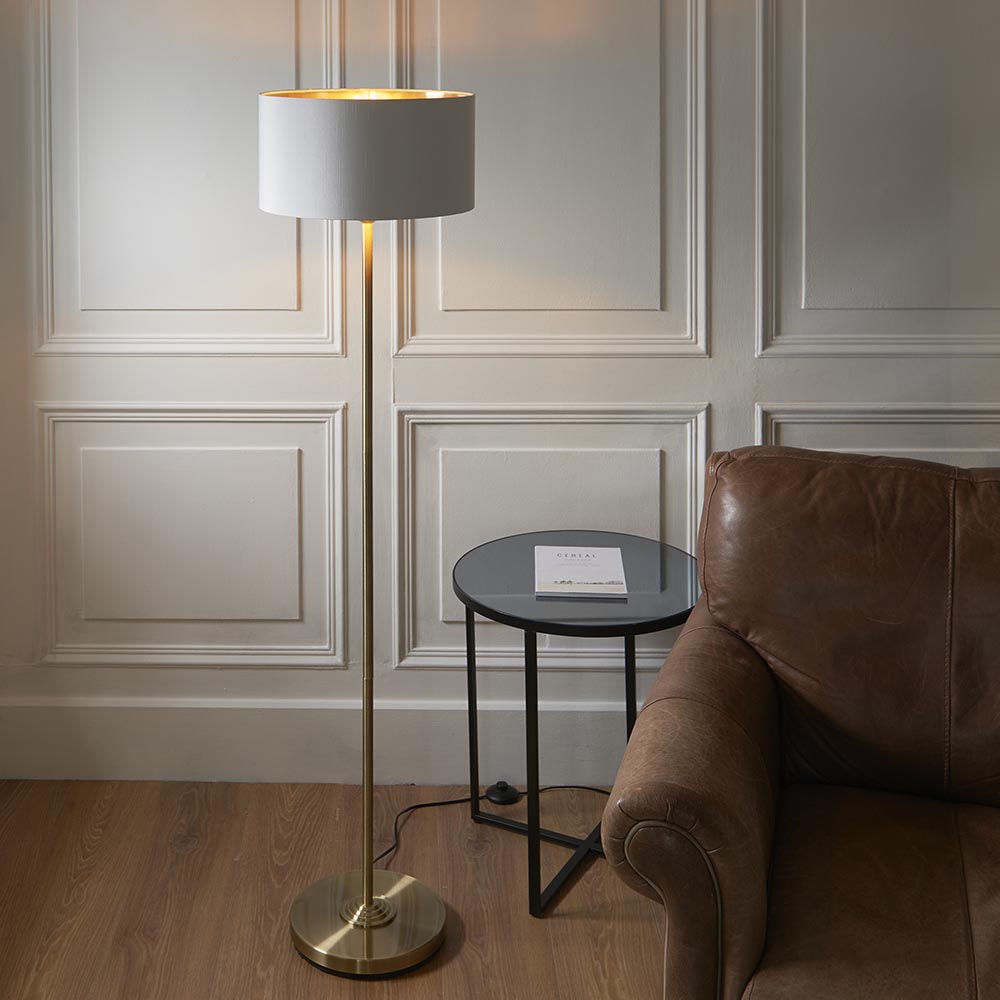 Endon Brenley floor lamp with brass stem, round base and white fabric shade with gold inner, lit beside a leather sofa and black side table.