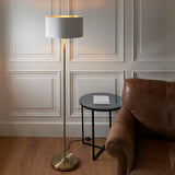 Endon Brenley floor lamp with brass stem, round base and white fabric shade with gold inner, lit beside a leather sofa and black side table.