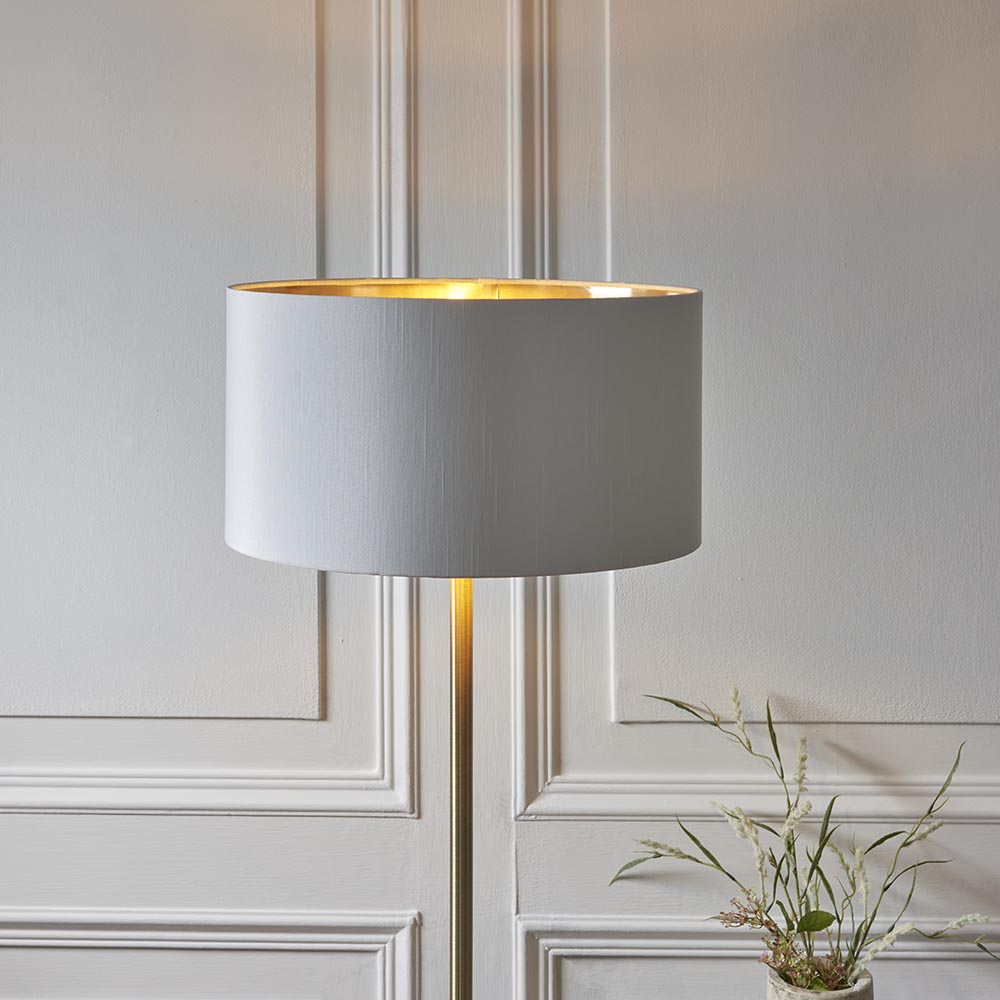 Endon Brenley floor lamp with slim brass stem and white faux silk drum shade lined in gold, lit beside a pale panelled wall.