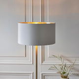 Endon Brenley floor lamp with slim brass stem and white faux silk drum shade lined in gold, lit beside a pale panelled wall.