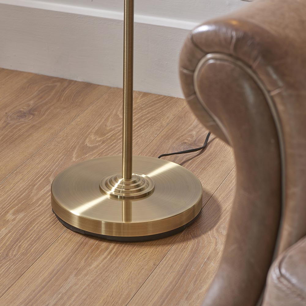 Close-up of the Endon Brenley floor lamp's circular brushed brass base and slim stem on wood flooring beside a brown leather armchair.