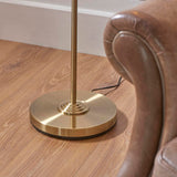 Close-up of the Endon Brenley floor lamp's circular brushed brass base and slim stem on wood flooring beside a brown leather armchair.