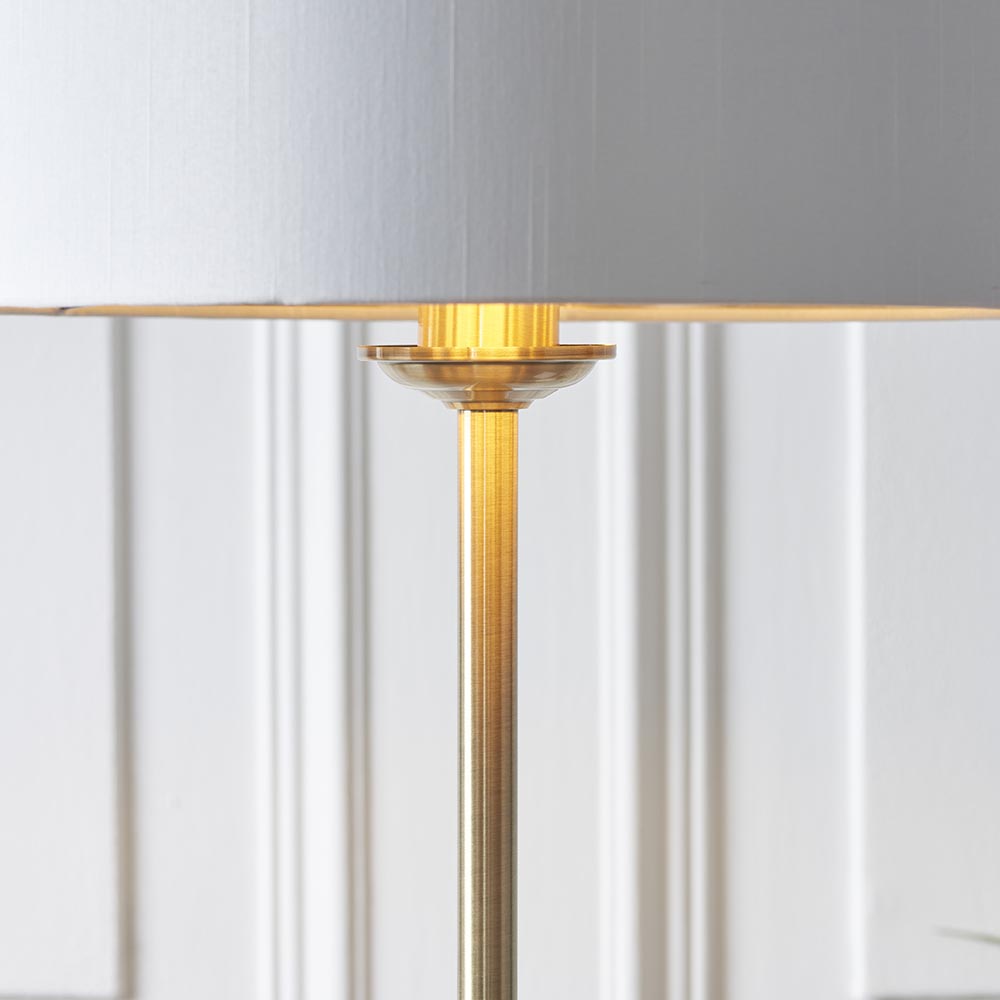 Close-up of the Endon Brenley floor lamp's brushed brass stem and shade fitter, with a white fabric shade above and panelled wall behind.