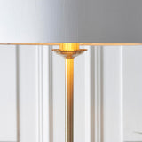 Close-up of the Endon Brenley floor lamp's brushed brass stem and shade fitter, with a white fabric shade above and panelled wall behind.