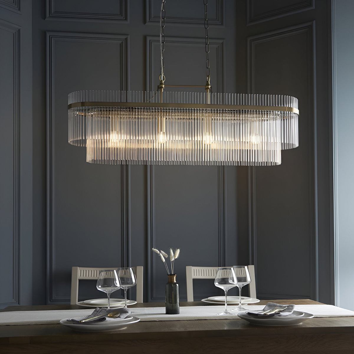 Beaumont 4 Light Linear Pendant in antique brass, with clear glass rods along an oval bar, hung on chains above a dining table in a dark panelled room.