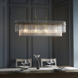 Beaumont 4 Light Linear Pendant in antique brass, with clear glass rods along an oval bar, hung on chains above a dining table in a dark panelled room.