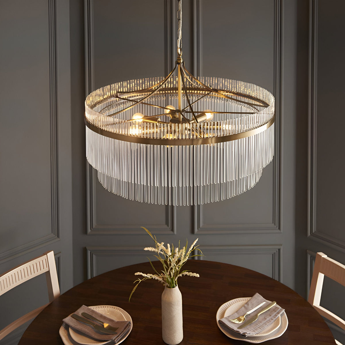 Endon Beaumont 7 Light Pendant in antique brass with tiered clear glass rods, hanging above a round wooden dining table against a grey panelled wall.