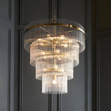Beaumont 16 Light Chandelier in antique brass, a large tiered fitting with clear glass rods and exposed bulbs, hung against a dark panelled wall.