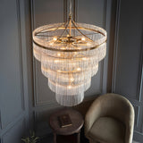 Beaumont 16 light tiered chandelier in antique brass with clear glass rods, hung in a room with panelled grey walls, armchair and side table below.