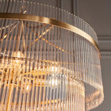 Close-up of the Beaumont 16 Light Chandelier in antique brass, showing tiered clear glass rods and exposed bulbs around a curved brass band.