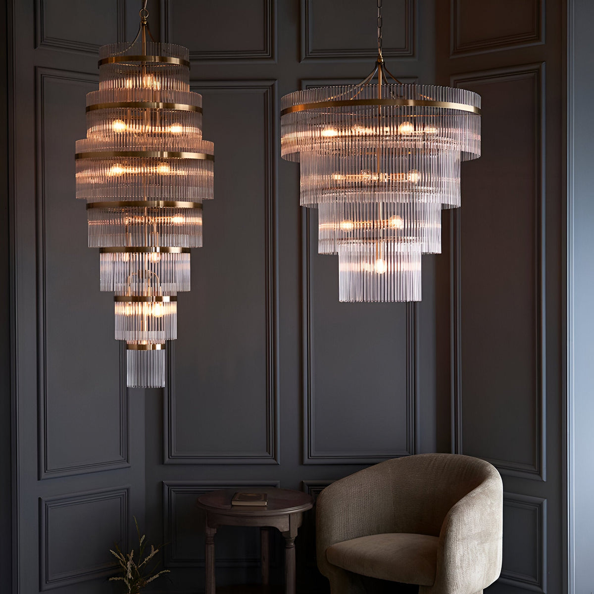 Beaumont 16 Light Chandelier in antique brass with tiered clear glass rods, shown alongside a smaller matching pendant in a dark panelled room with an armchair.