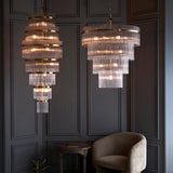 Beaumont 16 Light Chandelier in antique brass with tiered clear glass rods, shown alongside a smaller matching pendant in a dark panelled room with an armchair.