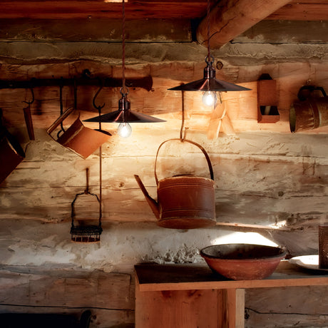 The rustic wooden interior highlights industrial design elements with the Sienna 1 Light Pendant in copper. Various vintage metal containers, such as a watering can, are mounted on the wall. A wooden shelf supports a shallow bowl, while soft light casts warm shadows throughout the space.