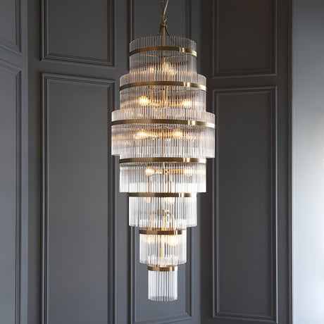 Endon Beaumont 21 light chandelier in antique brass with multi-tiered clear glass rods, hung from a chain against a grey panelled wall.