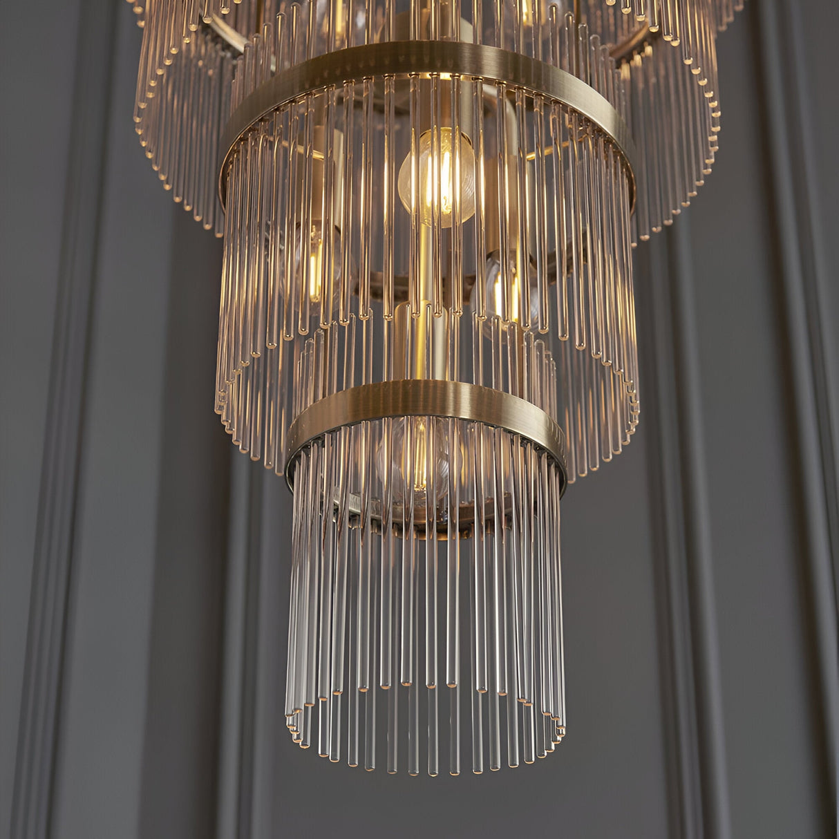 Close-up of Endon Beaumont 21 Light Chandelier in antique brass with tiered clear glass rods and lit E14 bulbs against a grey panelled wall.