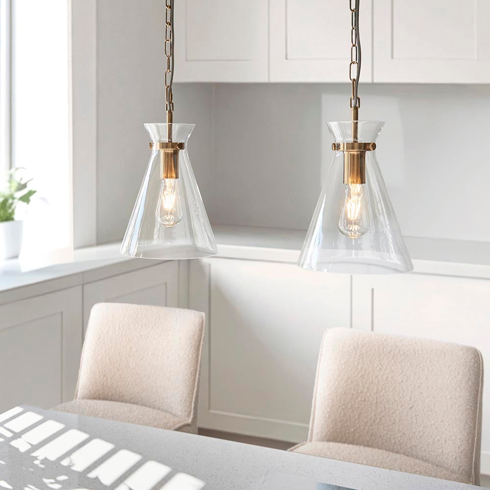 Endon Brenbury pendant lights in brushed brass with clear tapered glass shades and exposed E27 bulbs, hung on chains above a kitchen dining area.