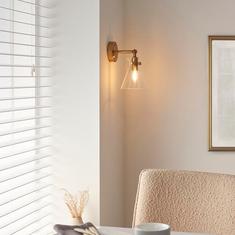Endon Brenbury antique brass wall light with adjustable knuckle and clear glass cone shade, lit above a boucci chair beside a window with white blinds.