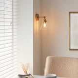 Endon Brenbury antique brass wall light with adjustable knuckle and clear glass cone shade, lit above a boucci chair beside a window with white blinds.