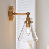 Endon Brenbury wall light in antique brushed brass with adjustable knuckle, clear glass conical shade and exposed E14 bulb, mounted beside a window.
