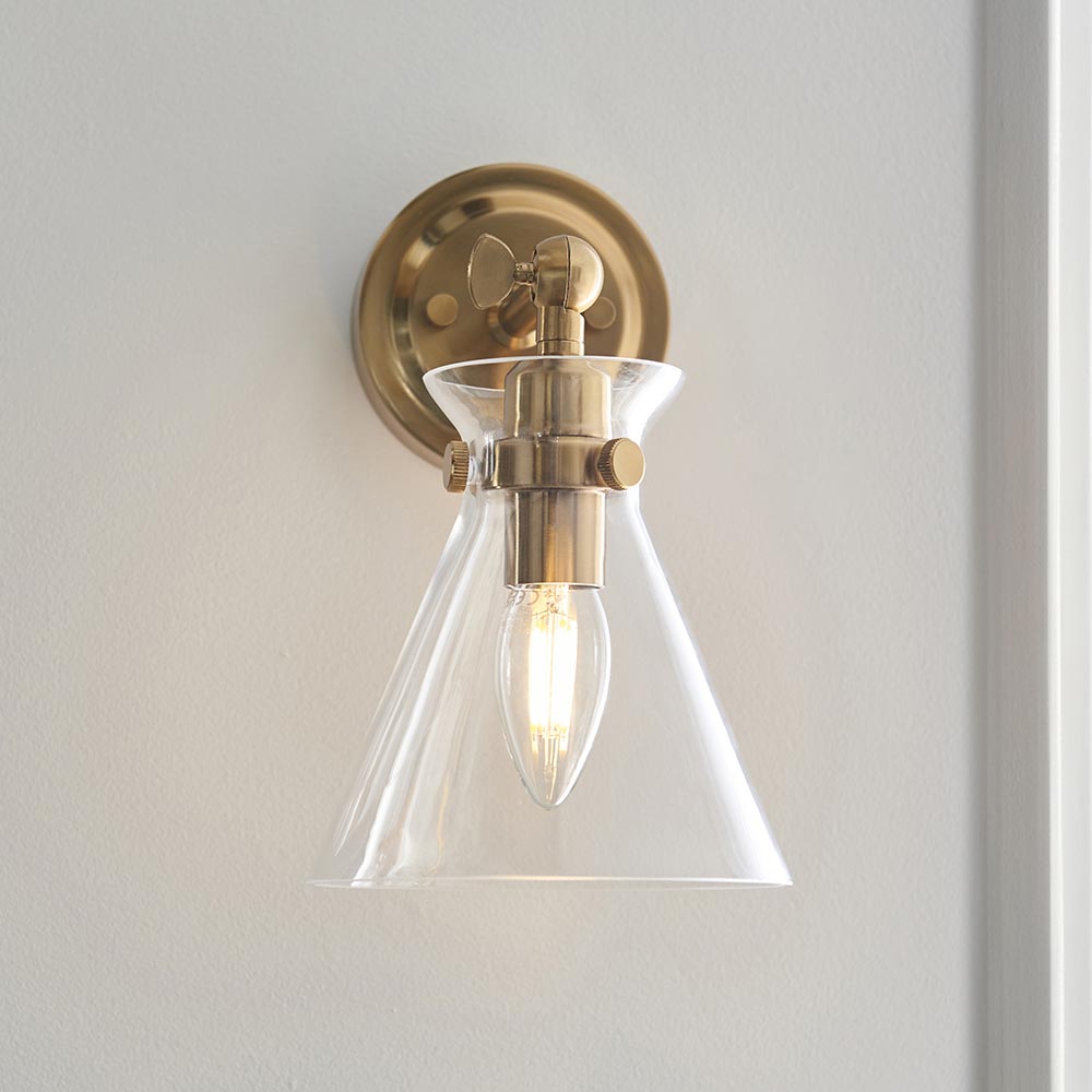 Endon Brenbury wall light with brushed antique brass fitting, adjustable knuckle and clear glass cone shade around an exposed E14 bulb, on a pale wall.