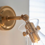 Close-up of the Endon Brenbury wall light showing its brushed antique brass adjustable knuckle, round backplate and clear glass shade.