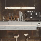 Bowie LED Alabaster Linear Pendant Light hanging above a café-style counter, featuring a long sectioned alabaster cylinder with warm white illumination over a stone countertop.