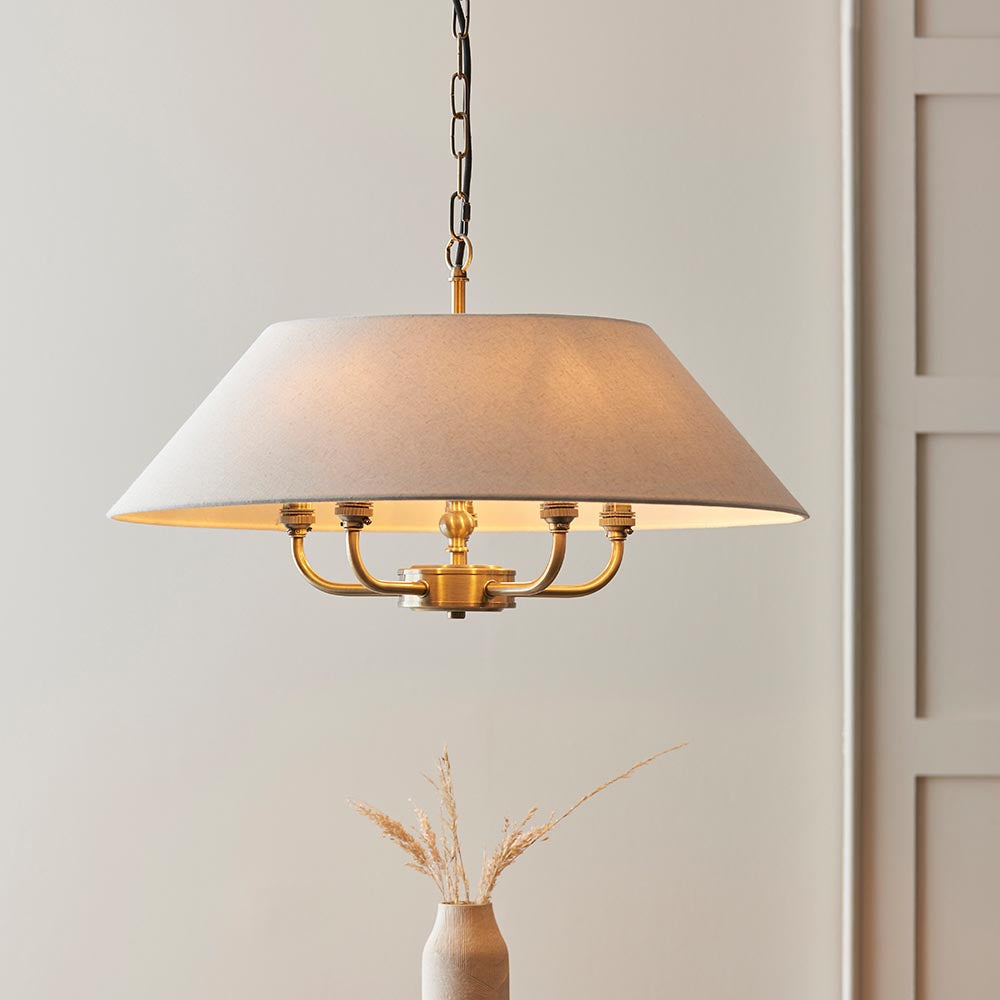Caldmere 5 Light Pendant in brass with a wide natural fabric drum shade, suspended from a chain, shown in a room setting with a vase nearby.