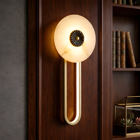 The Glintara LED Radiant Disc Alabaster Wall Light in Soft White & Brass is mounted on a wood-paneled wall next to bookshelves filled with books and a glass terrarium with succulents.