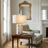 A cozy sitting area with a light green armchair and ottoman is next to a wooden table holding the Helvarn 1 Light Framed Block Marble Table Lamp - Brass & White, a tea set, and book, with a gold-framed mirror and fireplace adding elegance to the bright room.
