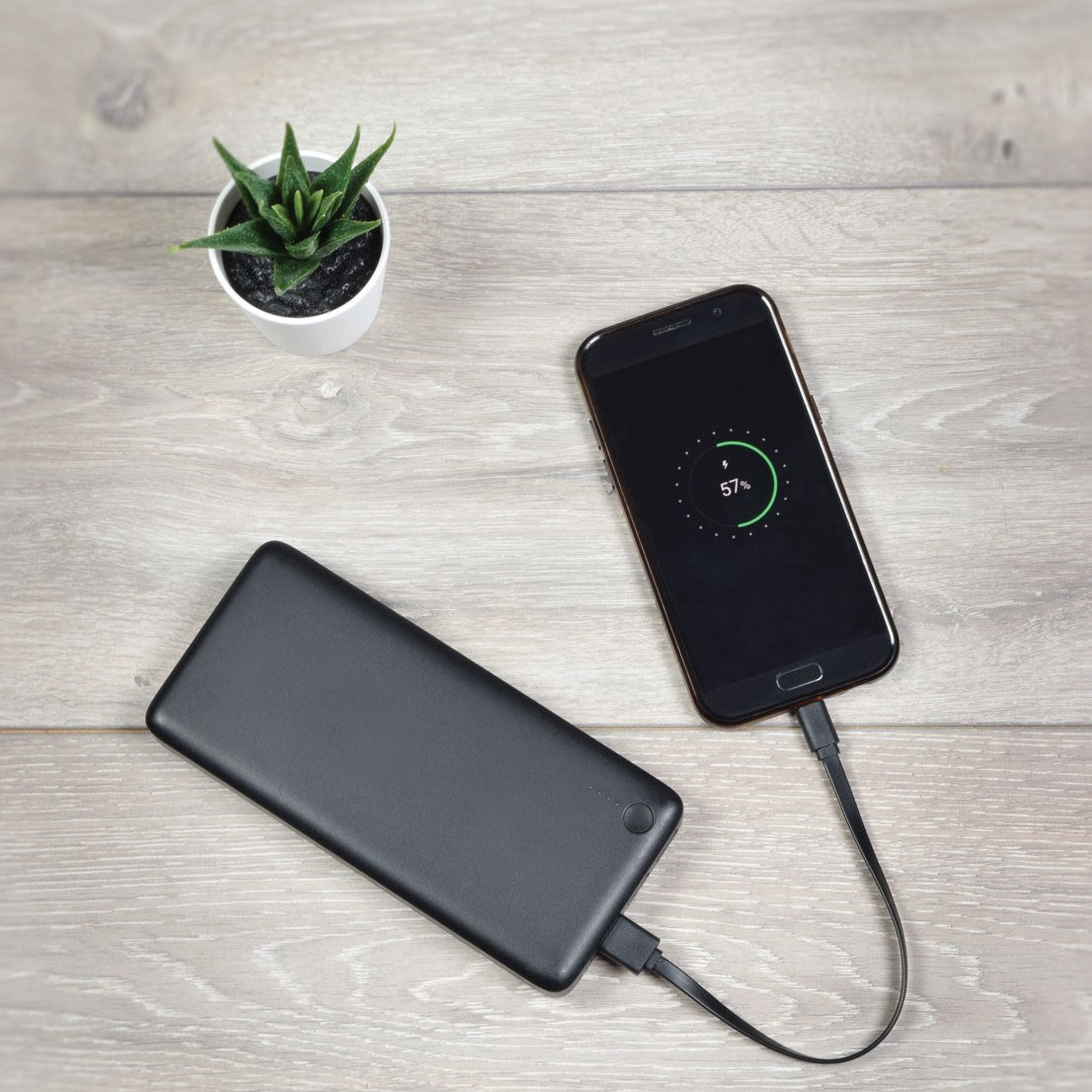 Black power bank charging a smartphone at 57% via AV:Link USB Type-A to C flat cable on a wooden surface beside a small potted succulent.