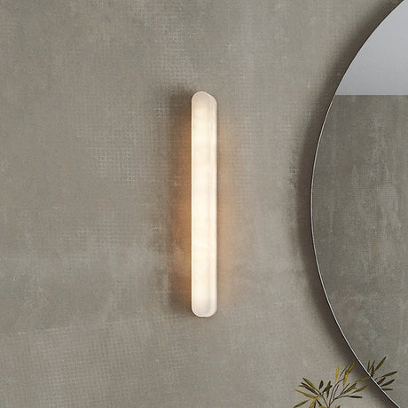 Bastian LED Alabaster Linear Wall Light, a slim vertical alabaster bar with rounded ends glowing warm white, mounted on a textured grey wall beside a round mirror.