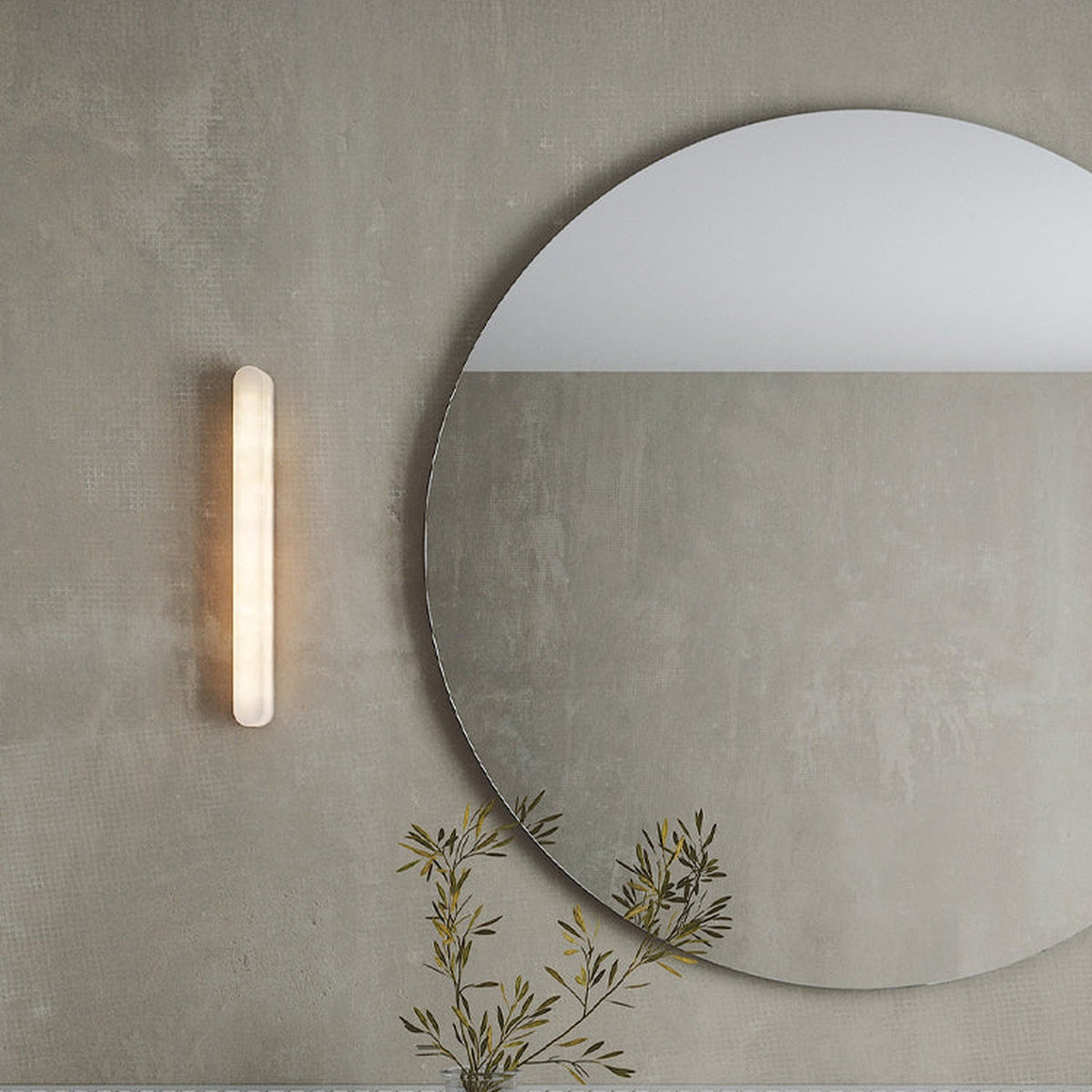 Bastian LED Alabaster Linear Wall Light, a slim vertical bar with rounded ends and warm inner glow, mounted beside a round mirror on a grey wall.