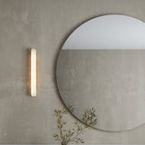 Bastian LED Alabaster Linear Wall Light, a slim vertical bar with rounded ends and warm inner glow, mounted beside a round mirror on a grey wall.