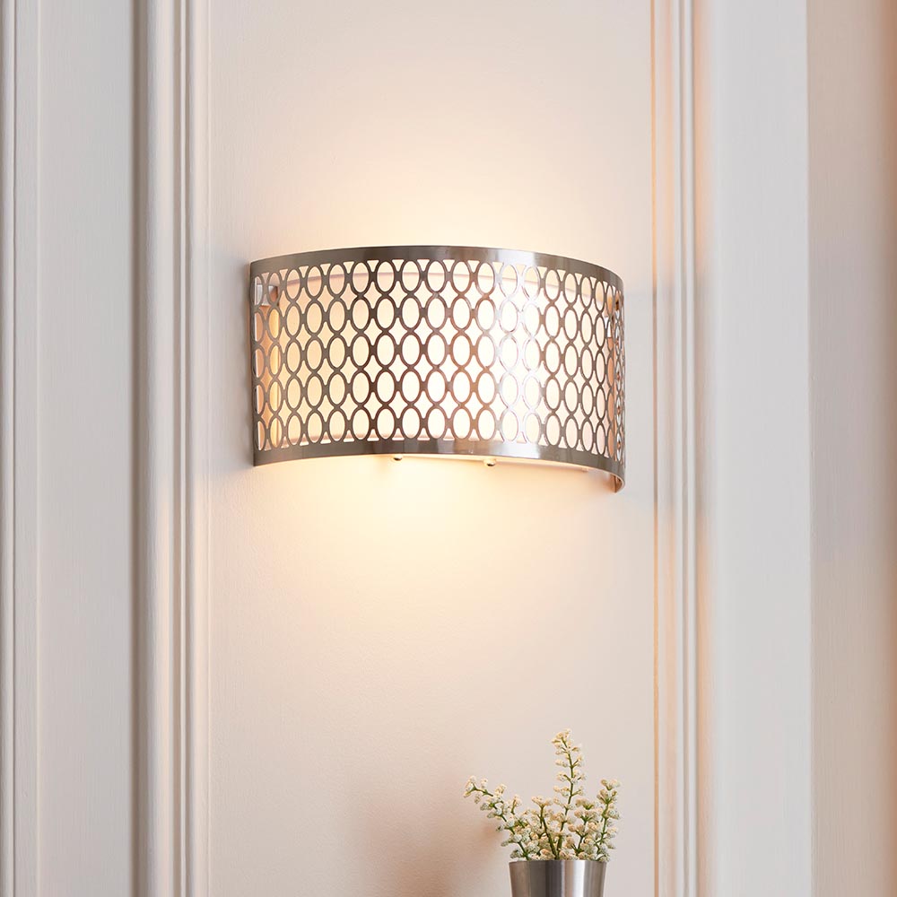 Endon Rosaline 2 light wall light in satin nickel with geometric fretwork, white fabric inner shade and frosted glass diffusers, lit on a panelled wall.