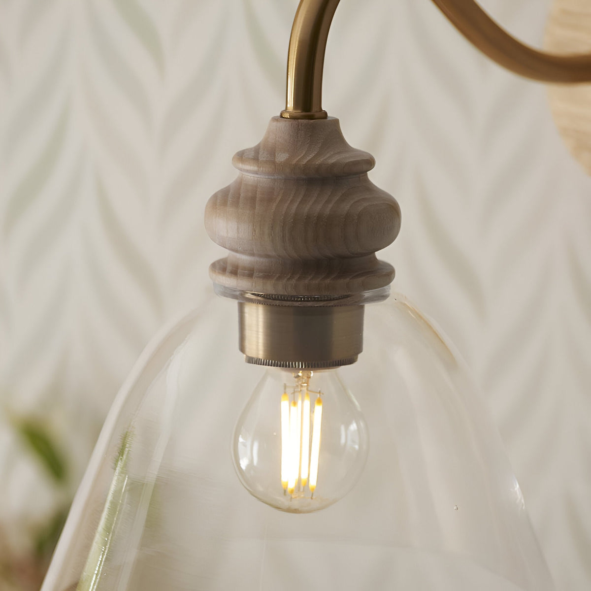 Close-up of Endon Bevwick wall light showing turned natural ash detail, aged brass arm, clear flared glass shade and lit E14 filament bulb.