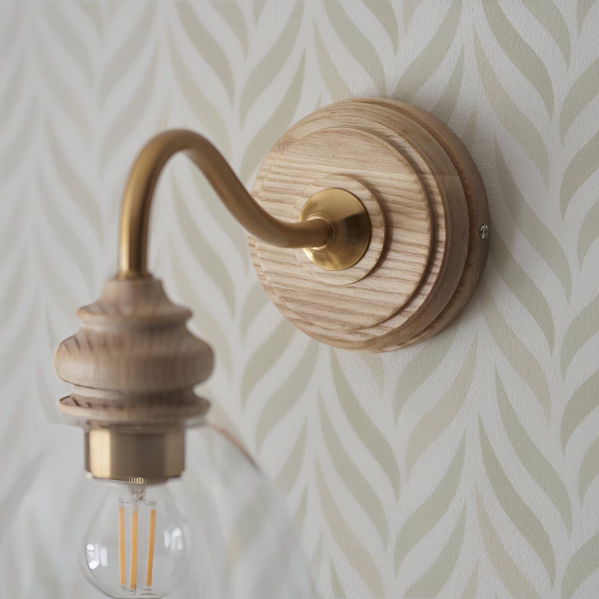 Close-up of Endon Bevwick wall light showing turned natural ash backplate, aged brass swan-neck arm and E14 lampholder against leaf-patterned wallpaper.
