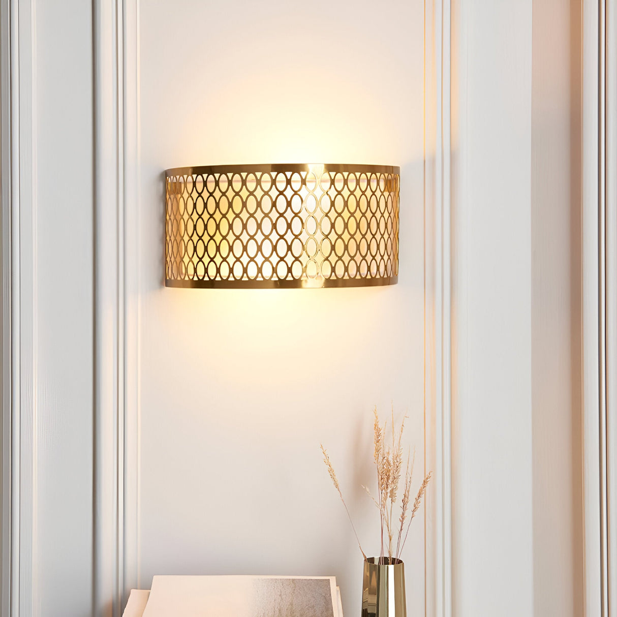 Rosaline 2-light wall light in gold with curved geometric fretwork, illuminated on a wall between panelled mouldings above a vase of dried grasses.