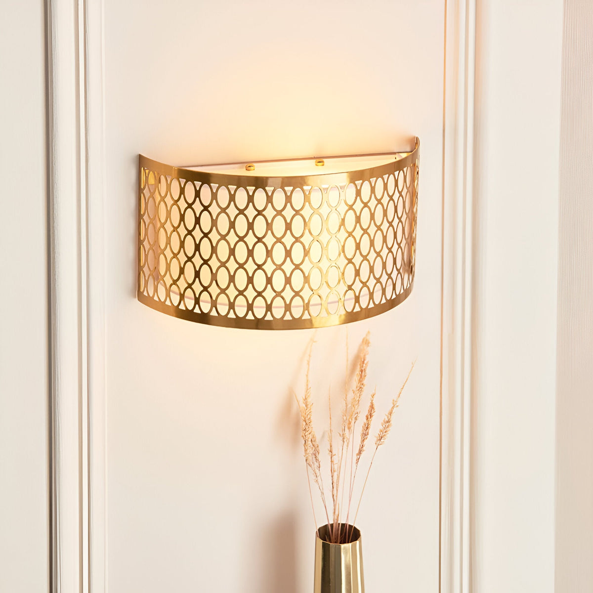 Rosaline 2 Light Wall Light in gold with geometric fretwork outer and white fabric inner shade, mounted on a wall beside a vase with dried grasses.