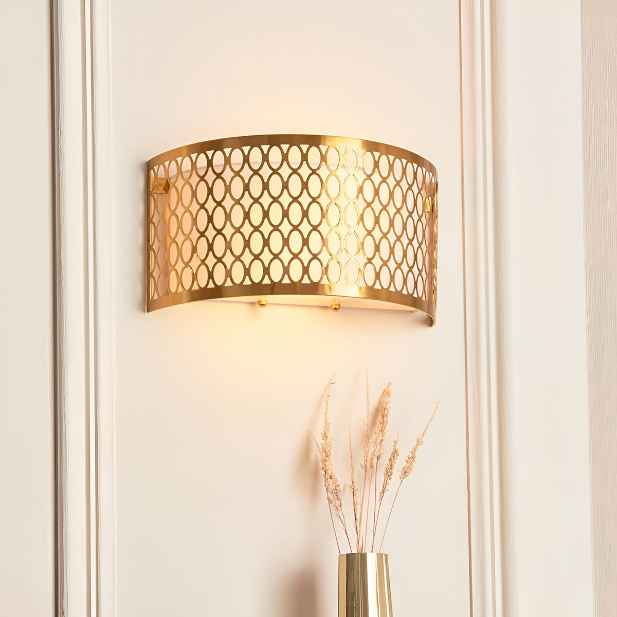 Rosaline 2 Light Wall Light in gold with curved geometric metal fretwork and white diffuser, illuminated on a panelled wall above a dried floral arrangement.