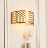 Rosaline 2 Light Wall Light in gold with curved geometric metal fretwork and white diffuser, illuminated on a panelled wall above a dried floral arrangement.